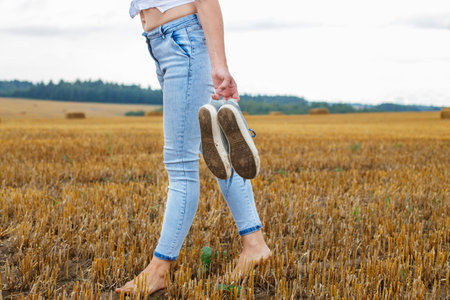 barefoot girl with sneakers in hand walking in the agricultural field with haystack and bales after harvesting.の写真素材