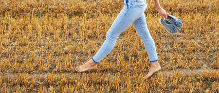 barefoot girl with sneakers in hand walking in the agricultural field with haystack and bales after harvesting.の写真素材