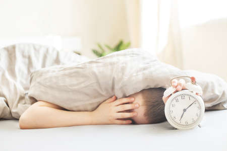 boy lying on the bed under a blanket and stopping alarm clock in the morning. childs hand reaching for the alarm clock to turn it off.の写真素材