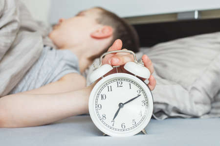 boy lying on the bed and stopping alarm clock in the morning. childs hand reaching for the alarm clock to turn it off.の写真素材