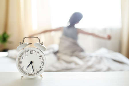 little boy sitting and stretching in the bed at home in the morning on a window background with alarm clockの写真素材
