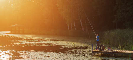 Little boy standing and fishing on a wooden dock at the sunset. bannerの写真素材