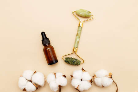 Massage quartz roller for the face of natural nephritis with serum oil in brown glass bottle with a flower of cotton on beige background.の写真素材