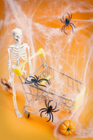 skeleton with a pumpkin near the trolley, spider webs and spiders on an orange background. Halloween shopping concept.の写真素材
