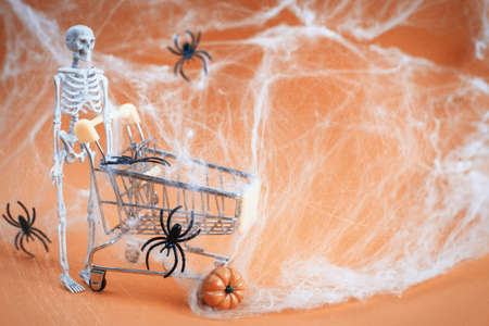 skeleton with a pumpkin near the trolley, spider webs and spiders on an orange background. Halloween shopping concept.の写真素材