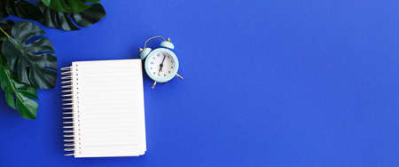 Top view of blank note paper, alarm clock and monstera leaf on blue background. bannerの写真素材