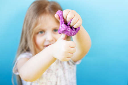 Blonde girl hold purple glitter slime and make thumbs up sign isolated on a blue background. child playing with a slime toy. Making slime. Copy space. DIY concept.の写真素材