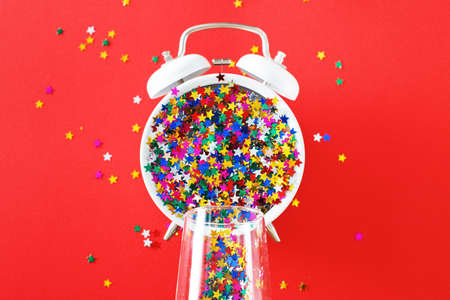A glass with multicolored sequins, alarm clock and star shaped confettie on the red background. Party, new year, celebration conceptの写真素材