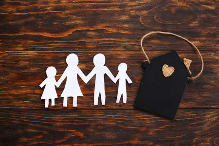 Happy paper cut family holding hands on wooden background near little toy house.の写真素材