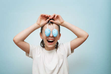 young smiling boy holding Easter eggs in his hand and covering his eyes with it on a blue...