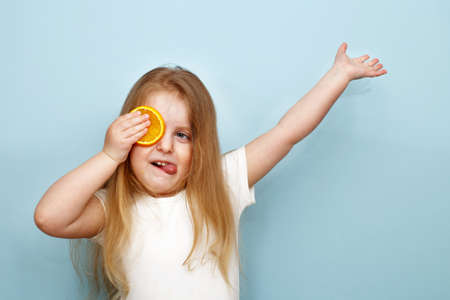 little happy girl with halves of oranges covering eyes on a blue background. child having fun and showing tongue.の写真素材