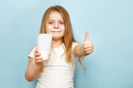 little beautiful girl holding milk in a glass on a blue background. baby drinks yogurtの写真素材