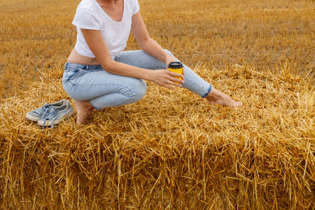 barefoot girl with sneakers and cardboard cup with coffee in hand standing in the agricultural field with haystack and bales after harvesting.の写真素材