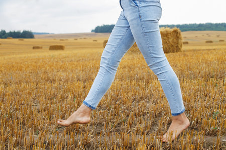 barefoot girl with sneakers in hand walking in the agricultural field with haystack and bales after harvesting.の写真素材