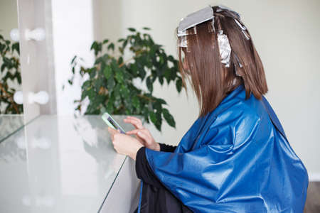 brunette girl dyes strands of hair in the salon and looks at his phone while waitingの写真素材