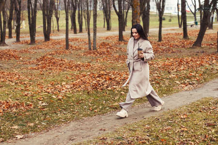 woman in a gray coat walking in the autumn park with a cup of hot drink.の写真素材