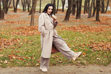woman in a gray coat walking in the autumn park with a cup of hot drink.の写真素材