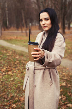 woman in a gray coat walking in the autumn park with a cup of hot drink.の写真素材