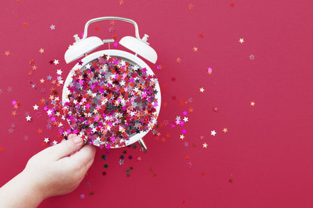 child's hands hold a white alarm clock with scattered sequins in the form of stars on a red...