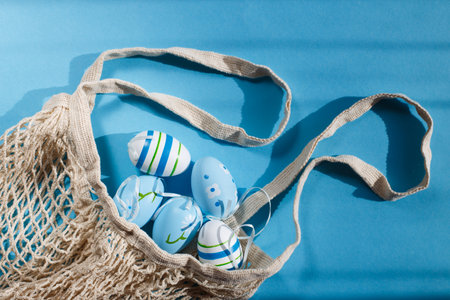 colorful handmade painted easter eggs in a string bag on blue background with hard shadows. minimal concept. Top view. An Easter card with a copy space for your textの写真素材