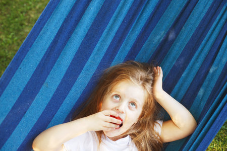 little blonde girl with long hair eats a strawberry in the garden lying in a hammock.の写真素材