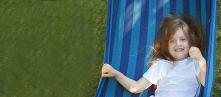portrait of a little cute girl with long hair resting on a hammock in the garden and smiling.の写真素材