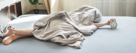 Sleeping boy lying on the bed under a blanket in his bedroom and stopping alarm clock on bedside table at home. Wake up in morning. Child reaching for alarm clock to switch it off. Bannerの写真素材