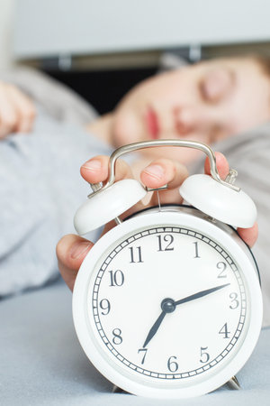 Sleeping boy lying on the bed in his bedroom and stopping alarm clock on bedside table at home. Wake up in morning. Child with closed eyes reaching for alarm clock to switch it off. Bannerの写真素材