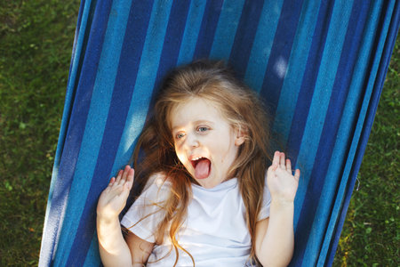 portrait of a little cute girl with long hair resting on a hammock in the garden and showing tongue.