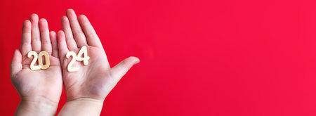 Child hands holding wooden number 2024 on palms on red background with copy space. Christmas greeting card, New Year, winter holidays sale and festive mood concept.の写真素材