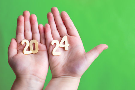 Child hands holding wooden number 2024 on palms on green background with copy space. Christmas greeting card, New Year, winter holidays sale and festive mood concept.の写真素材