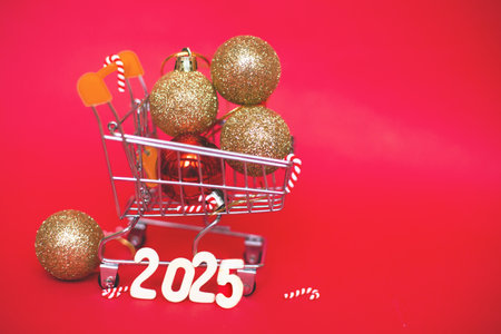 Christmas decoration tree toy balls in a small shopping trolley cart with garland and wooden numbers 2025 year on a red festive background. New Year, Winter holidays sale concept.の写真素材