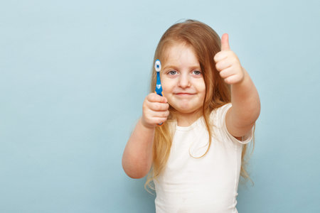 little happy girl holding her toothbrush and smiling on studio blue background showing thumb up sign. hygiene and healthy teeth concept.の写真素材