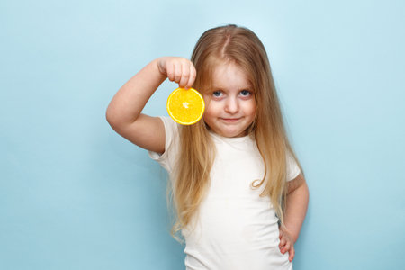 Portrait of happy little cute child girl holding halve of orange in hand and looking at camera on studio blue background. Kid eating vitamin C. Healthy nutrition concept.の写真素材