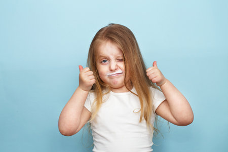 Portrait of cute little child girl looking at camera with dirty from drinking milk or kefir lips like mustache on studio blue background. Happy kid showing thumb up sign. Children health care conceptの写真素材