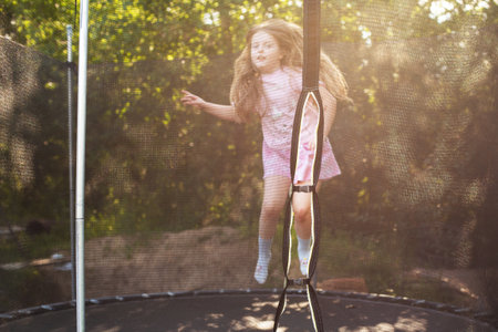 Child girl jumping on trampoline with net for safety. Happy smiling kid playing outdoors in the yard on summer vacation.の写真素材