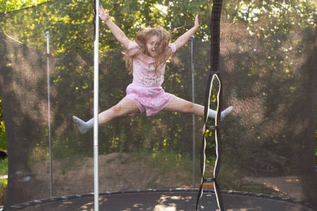 Child girl jumping on trampoline with net for safety. Happy smiling kid playing outdoors in the yard on summer vacation.の写真素材