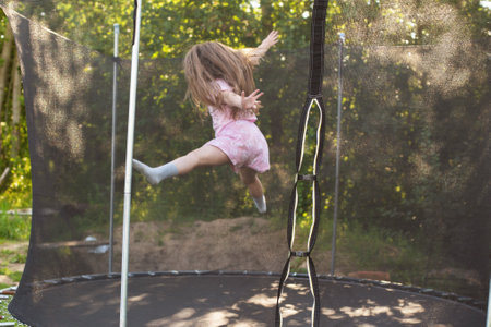 Child girl jumping on trampoline with net for safety. Happy smiling kid playing outdoors in the yard on summer vacation.の写真素材
