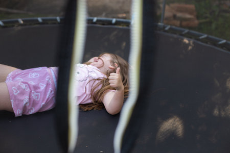 Portrait of a happy smiling excited child girl lying on trampoline outdoor and showing thumb up sign with joyful face expression. Kid playing on summer holiday. Active leisure concept.の写真素材
