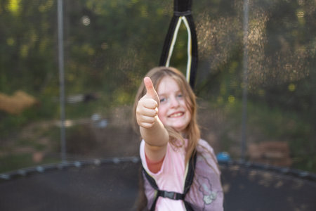Portrait of a happy smiling excited child girl sitting on trampoline outdoor and showing thumb up sign with joyful face expression. Kid playing on summer holiday. Active leisure concept.の写真素材