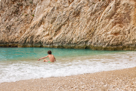 Young teenage boy tourist sitting in the shallow water of a beautiful beach with large cliff behind him. Child sitting at the seashore on summer holiday trip. Travel concept.の写真素材