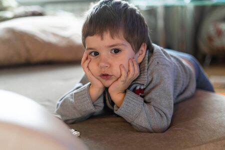 The boy lies on the sofa and propping his face with his hands