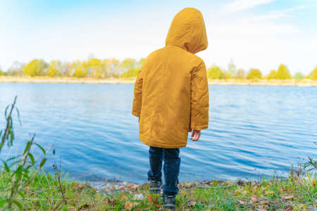 A boy in a yellow jacket stands near the lakeの写真素材