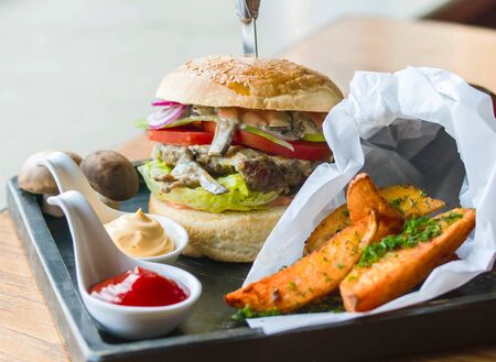 Burger with juicy meat cutlet and mushrooms, on a serving board, with sauces and fried potatoesの写真素材