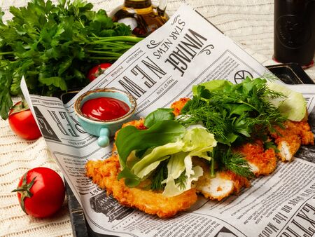 Deep-fried chicken breast with chopped herbs, on a serving paper in the style of a vintage newspaper, decorated with lettuce and spinach on a background of parsley and tomatoes. Ketchup, Olive Oil. Close-upのeditorial素材