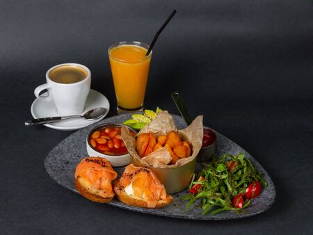 Breakfast on a beautiful plate - fried potatoes, beans, two sandwiches with fetax cheese and salmon, rucola and tomato salad with pesto, sliced avocado. On a dark background. On the left is a glass of orange juice with a straw and a cup of coffee.の写真素材