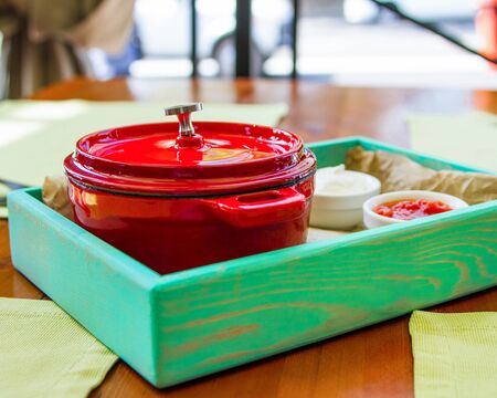 A red cast iron saucepan with a lid is served on a restaurant table with two sauces. The serving...
