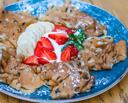 Pancakes with carved ornaments on a blue plate with cream, banana and strawberries. Close-upの写真素材