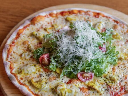 Caesar pizza on a wooden board with chicken and a slide of Caesar salad in the center, close-up. Fragment. Grated Parmesan Cheese. Lettuce leaves. Selective focus, side viewの写真素材