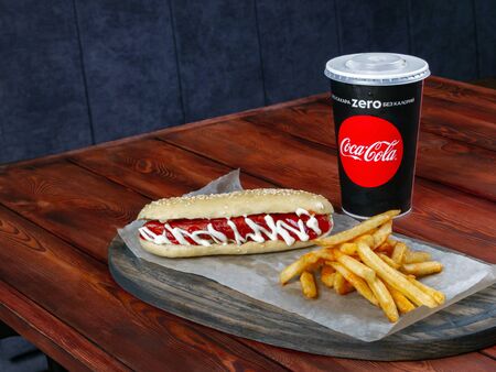 Hotdog decorated with sauces served with french fries and cola in a black paper cup. Fast food. Serving board. Wooden table. Copyspaceのeditorial素材